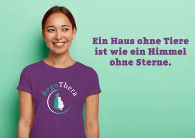 Eine lachende junge Frau trägt ein lilafarbenes T-Shirt. Auf dem T-Shirt ist das Logo von ReguThera aufgedruckt. Der Spruch "Ein Haus ohne Tiere ist wie ein Himmel ohne Sterne" ist auf dem grünen Hintergrund zu sehen.