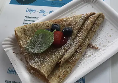 Ein Crêpes auf einem Pappteller bestreut mit Puderzucker. Darunter liegt die gestaltete Speisekarte des Weekender Blankenese.